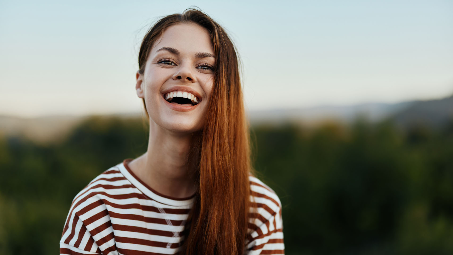 Young woman with long auburn hair laughing outdoors, wearing a striped top, with blurred green hills behind her
